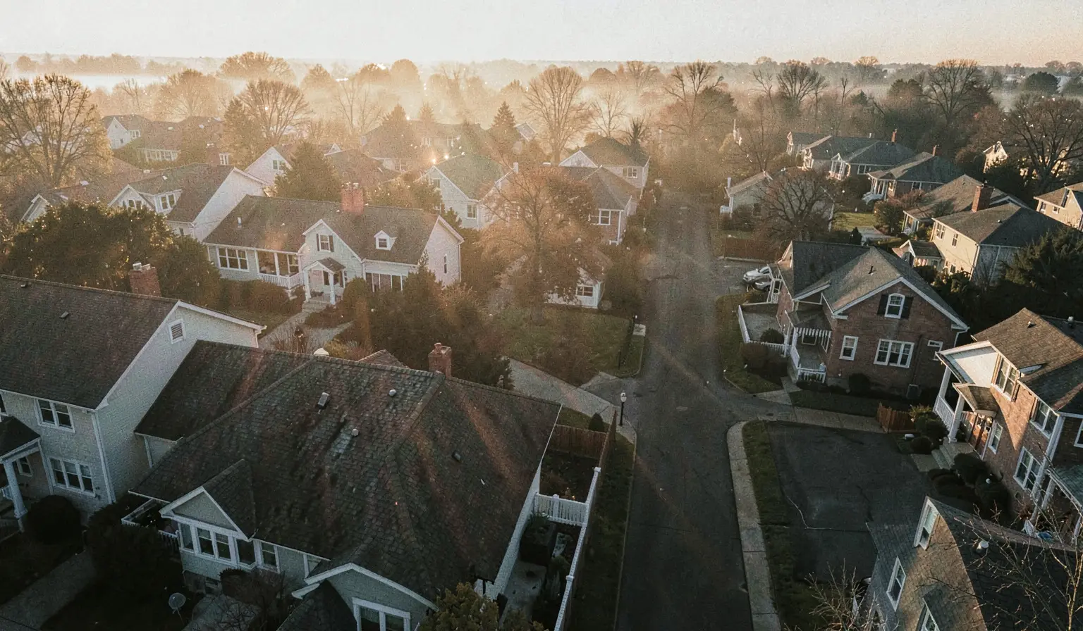 A neighbourhood of houses at dawn seen from above, mist lying over the treeline and a street running away toward the light